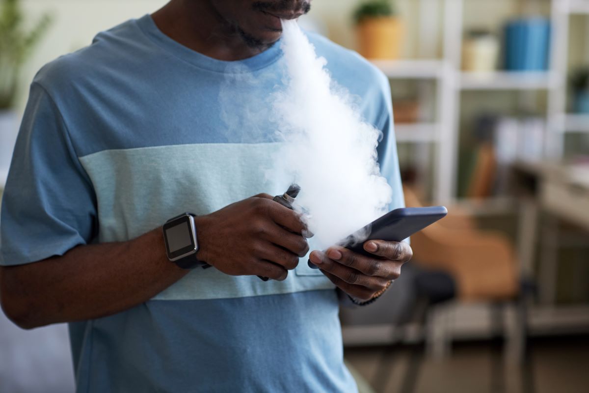 African American guy with e-cigarette in hand texting in smartphone and releasing clouds of white thick vapor from his mouth